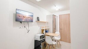 A small, modern room with a flat-screen TV mounted on the wall, a wooden door, a white table with two chairs, and a compact kitchenette with a microwave and refrigerator. - Good Deal Studio Room (No Kitchen) Bandaraya - Tallasa City Makassar Apartment (Makassar)