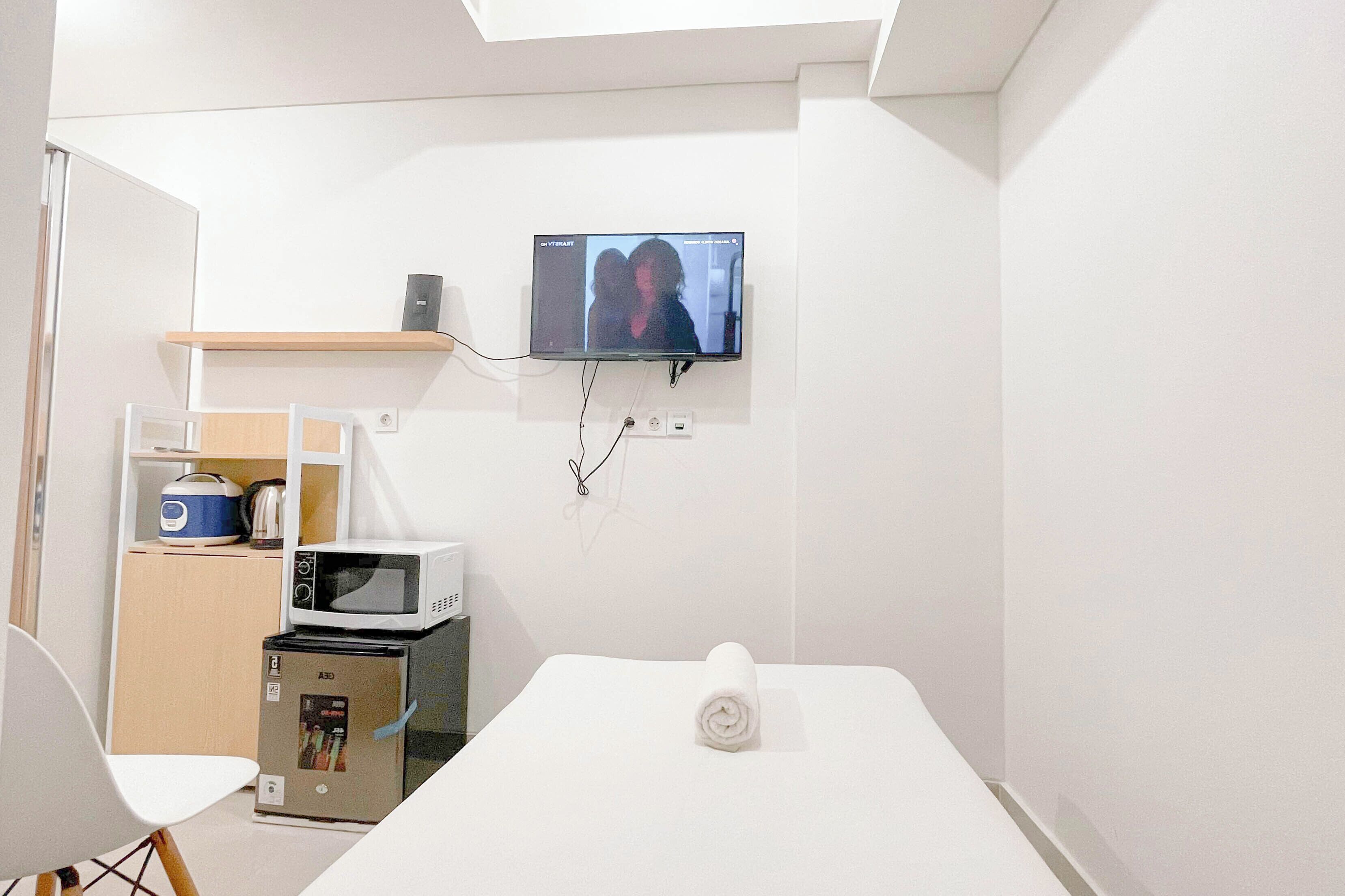 A compact room with a flat-screen TV mounted on the wall, a microwave, a small refrigerator, and a white table with a rolled towel.
