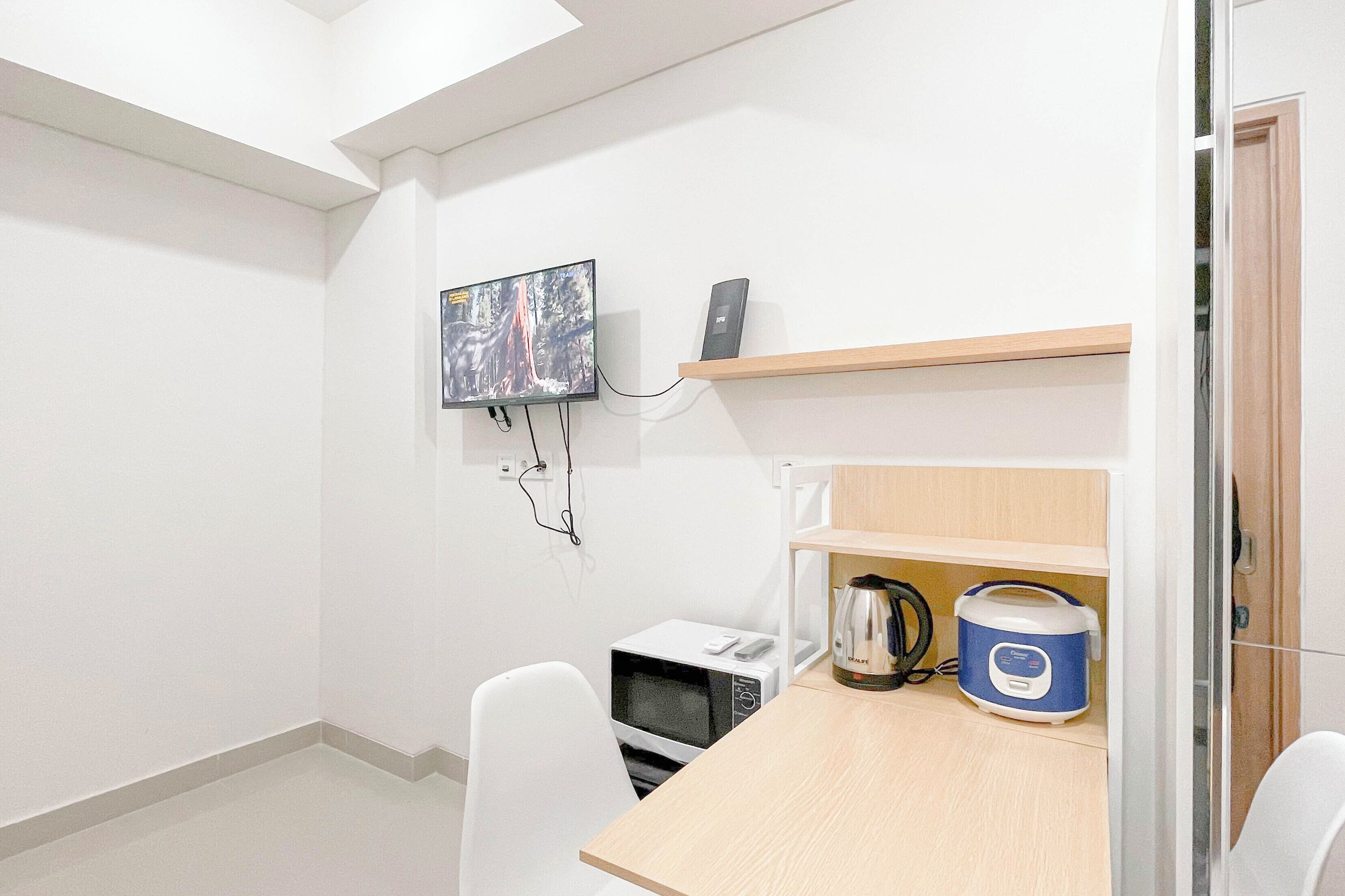 A small, modern room with a flat-screen TV mounted on the wall, a microwave, a kettle, and a rice cooker on a wooden table.