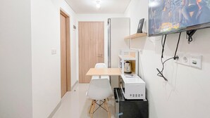 Een kleine, moderne kamer met een houten deur, een witte tafel met een stoel, een magnetron en een aan de muur bevestigde televisie.