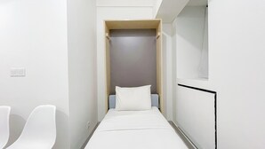 A narrow, modern hotel room with a single bed, a small seating area, and a wardrobe.