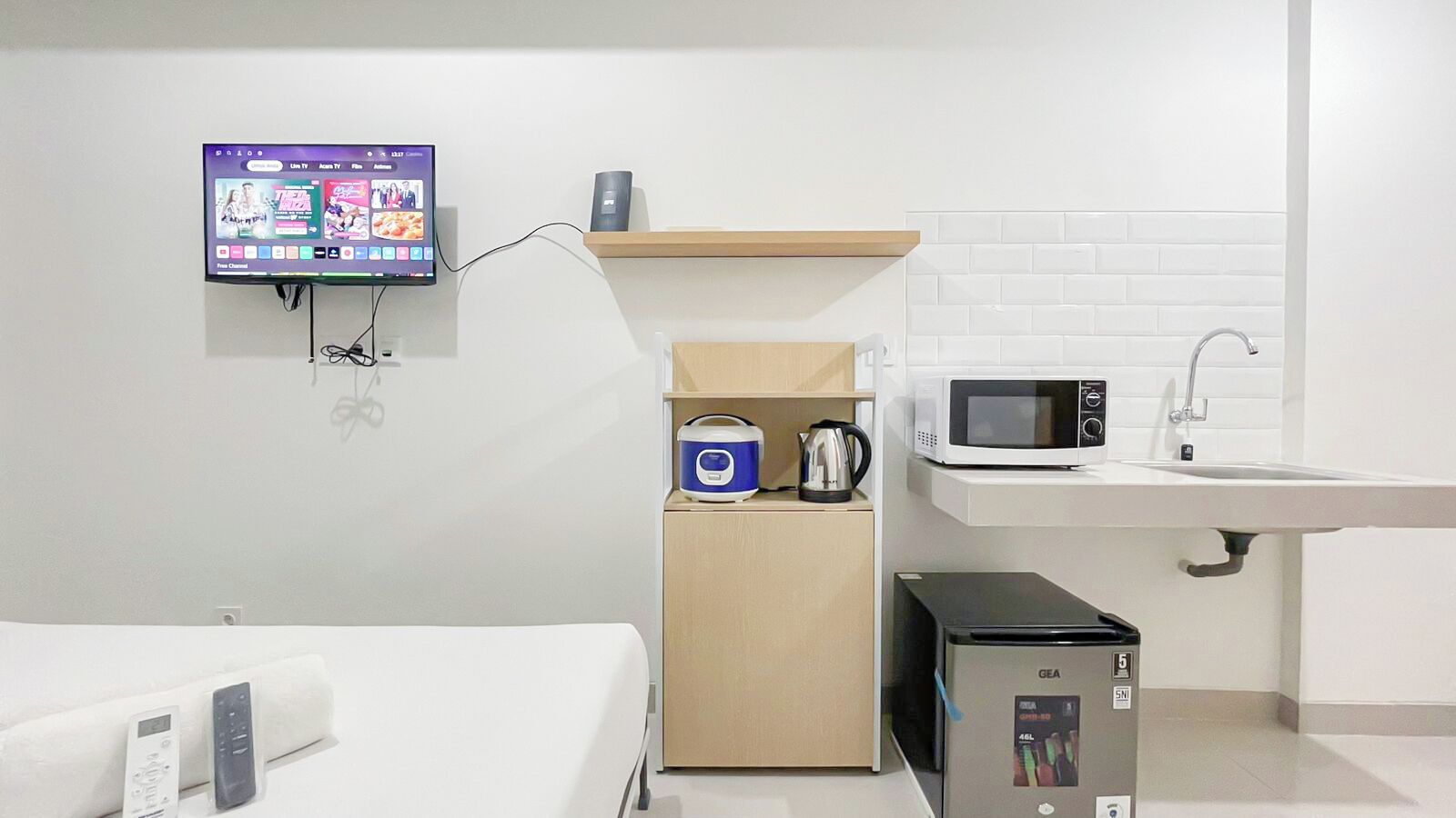 Quarto de hotel moderno com televisão de tela plana na parede, cozinha compacta com micro-ondas, chaleira e geladeira GEA, e uma pia branca.