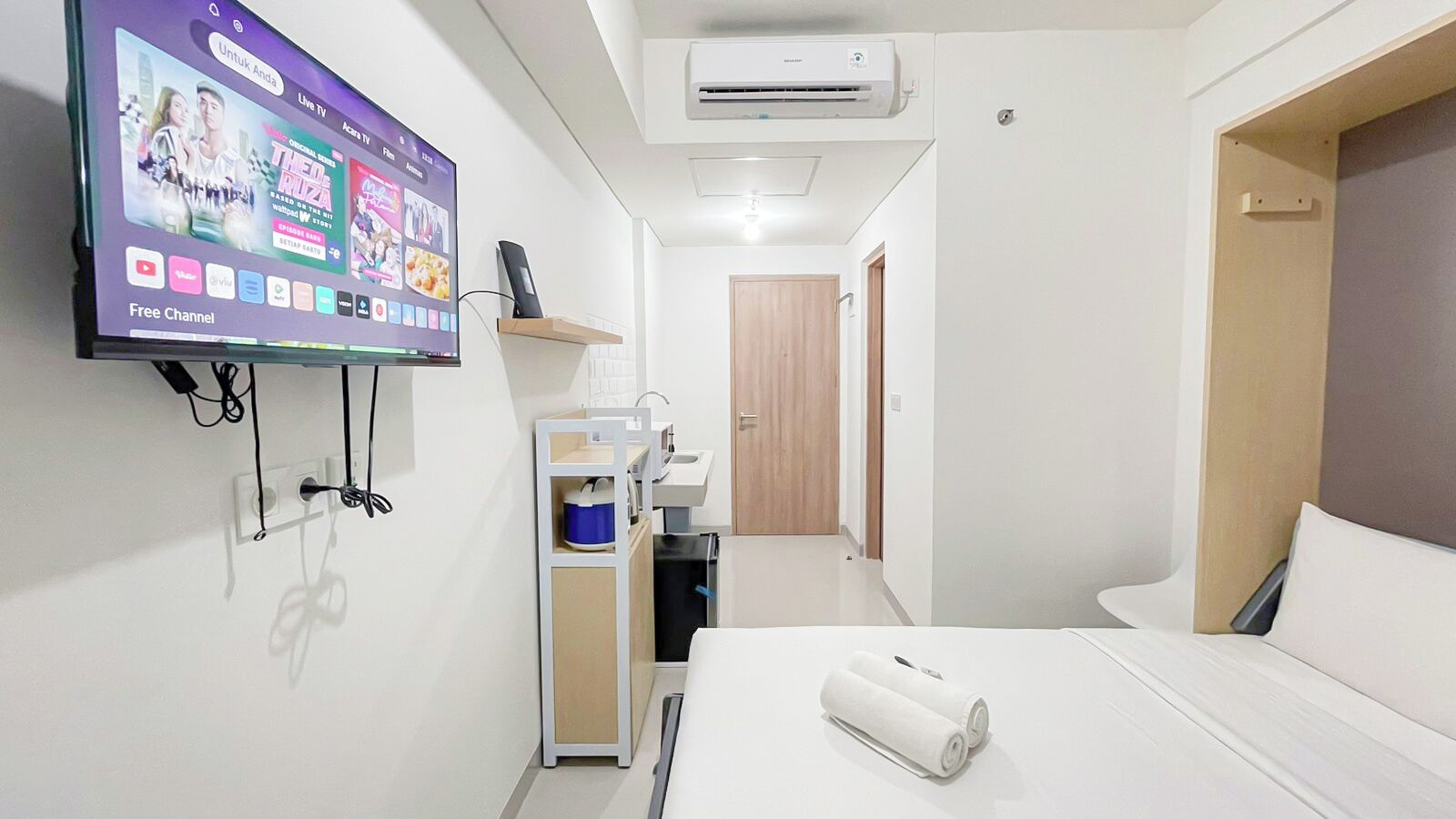 A hotel room with a bed, a TV mounted on the wall displaying various channels, a small kitchenette, and a door leading to another room.