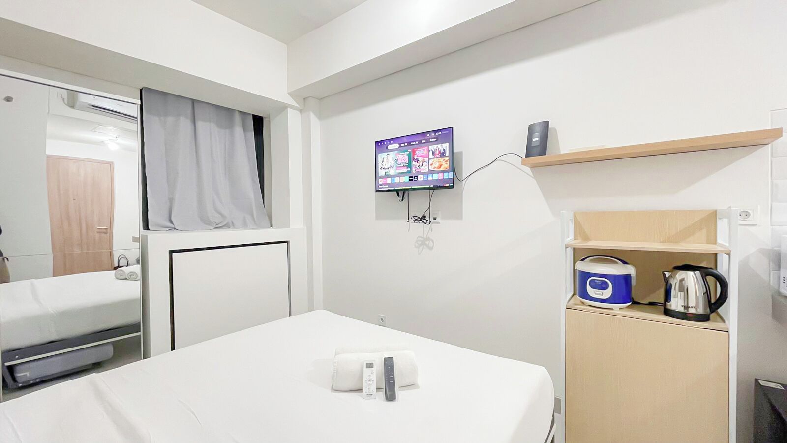 A hotel room with a bed, a TV mounted on the wall, a kettle, and a small shelf with a kettle.