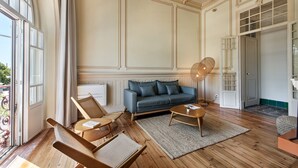 A spacious living room with a large window, a blue sofa, wooden flooring, and a decorative ceiling. - PG - 1C - T1 Superior - Palacete do Guadiana (Vila Real Santo Antonio)