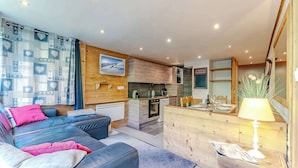 A modern living room with a sofa, a dining area with a table set for two, a kitchen with wooden cabinets, and a mounted picture of a snowy landscape.