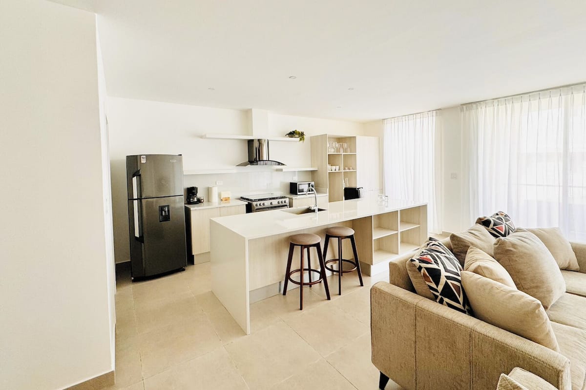 A modern kitchen with a central island, a refrigerator, and a dining area with a sofa and bar stools.