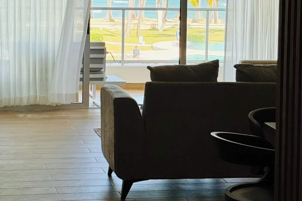 A room with a large window overlooking a beach and palm trees. There is a sofa and a small table in the room.
