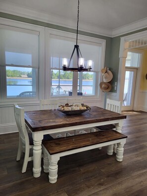 Un espace repas avec une table en bois, des chaises blanches, un lustre et une vue sur un lac, visible à travers de grandes fenêtres.