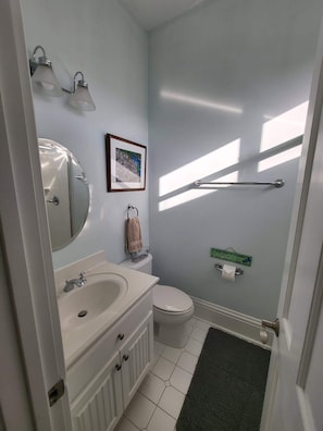 Une salle de bain avec un lavabo blanc, un miroir et des toilettes. On y trouve un porte-serviettes avec une serviette, un tableau encadré au mur et une pancarte sur la porte.