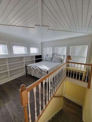 Une chambre à coucher comprenant un lit, des étagères intégrées, un escalier et de grandes fenêtres équipées de stores.