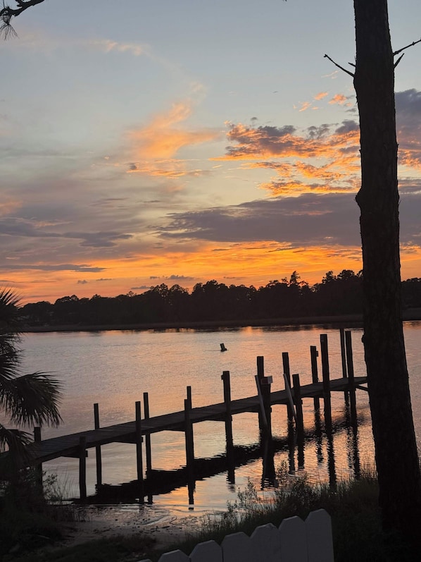 Un coucher de soleil sur un lac paisible, avec un quai en bois et des arbres au premier plan.