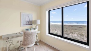 A room with a large window overlooking the beach, a chair, a desk with a lamp, and a framed picture on the wall. - Surf's Edge 305  Pet Friendly (Ocean City)