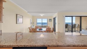 A kitchen with a granite countertop, dining area with chairs, and a view of the ocean through large windows. - Surf's Edge 305  Pet Friendly (Ocean City)