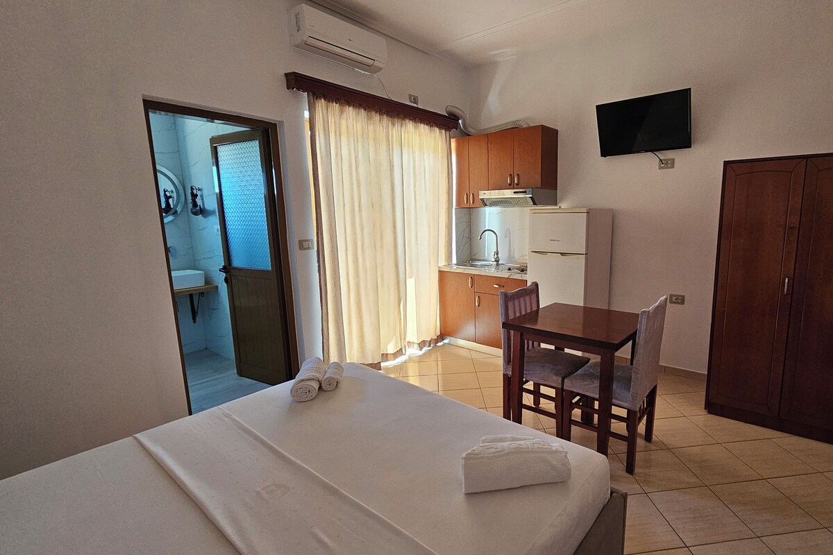 A hotel room with a bed, a desk, a chair, a TV, a wardrobe, a kitchen area, and a bathroom.