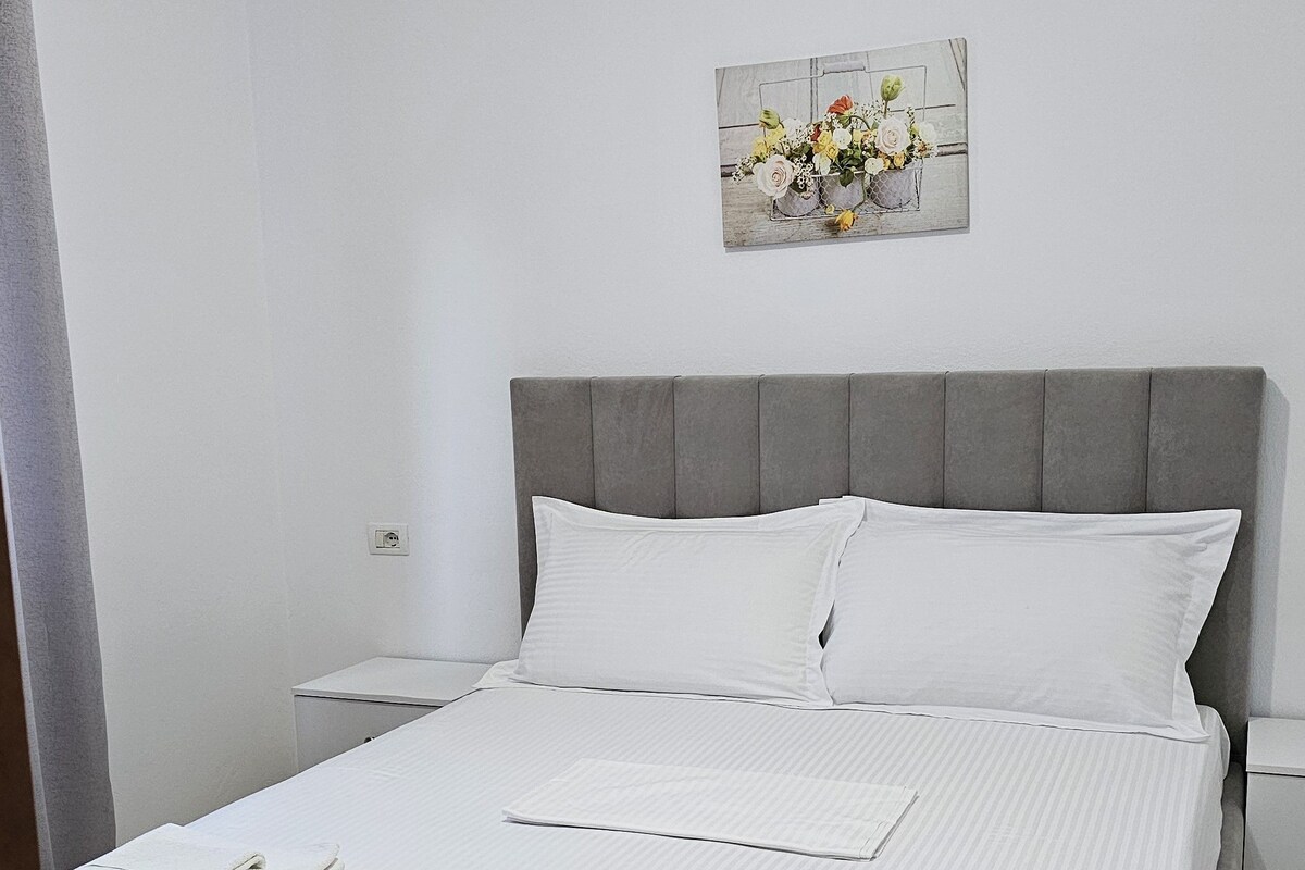 A neatly made bed with white linens and pillows, a grey upholstered headboard, and a small bedside table with a lamp.