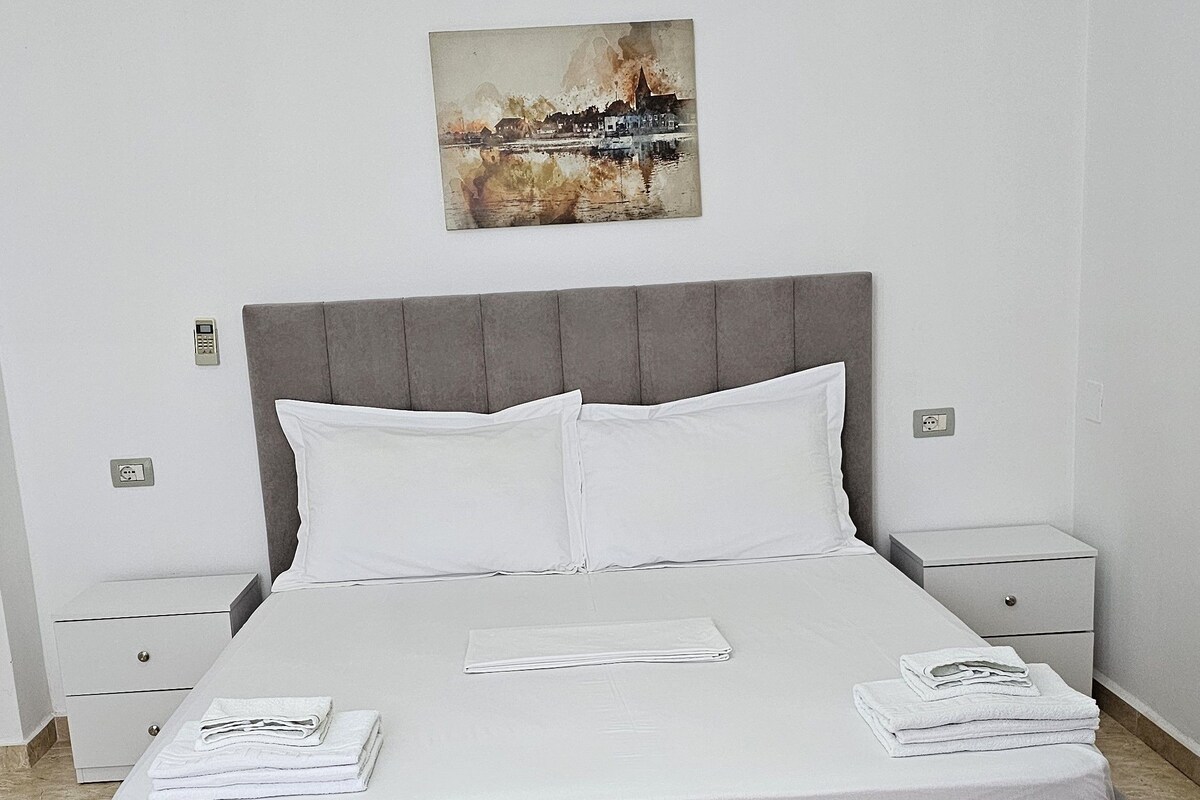 A neatly made bed with white linens, a grey headboard, and a painting above it. There are two bedside tables with lamps, a small wooden table in the foreground, and a tiled floor.