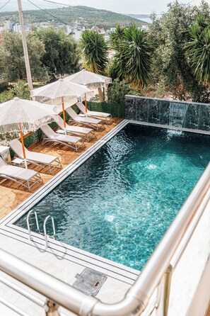 A rectangular pool with clear water, surrounded by lounge chairs and umbrellas, with a view of greenery and buildings in the background. - Vila Malo (Ksamil)
