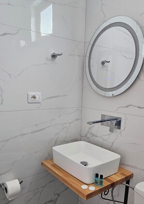A modern bathroom with a marble wall, a white sink, a mirror with a light, and a wooden shelf with toiletries. - Vila Malo (Ksamil)