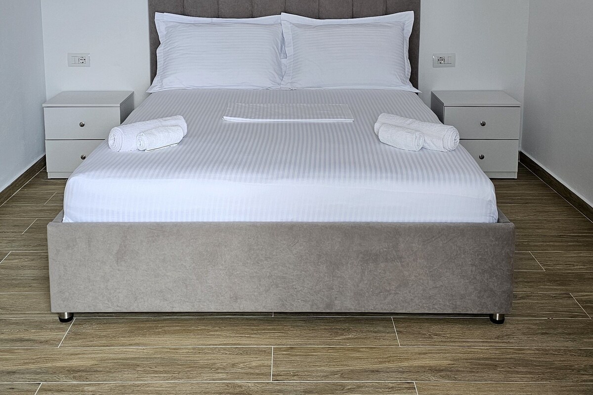 A neatly made bed with white linens and two pillows, a grey upholstered headboard, and a bedside table with a lamp.