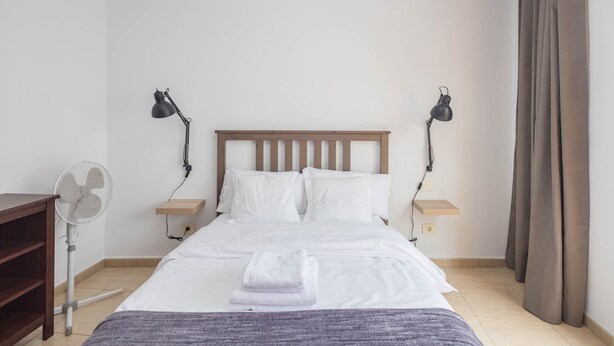 Ein ordentlich bezogenes Bett mit weißen Leinen, zwei Nachttische mit Lampen, ein Holzkopfende, ein kleiner Holznachttisch, ein Ventilator und ein gefaltetes Handtuch auf dem Bett.