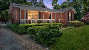 A brick house with a covered porch, surrounded by well-maintained hedges and a gravel driveway. - The Oleander Cottage (Wilmington)