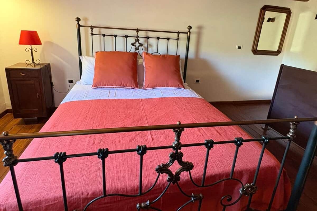 A bed with a metal frame, a red bedspread, and two orange pillows. There is a wooden floor, a nightstand with a lamp, and a mirror on the wall.