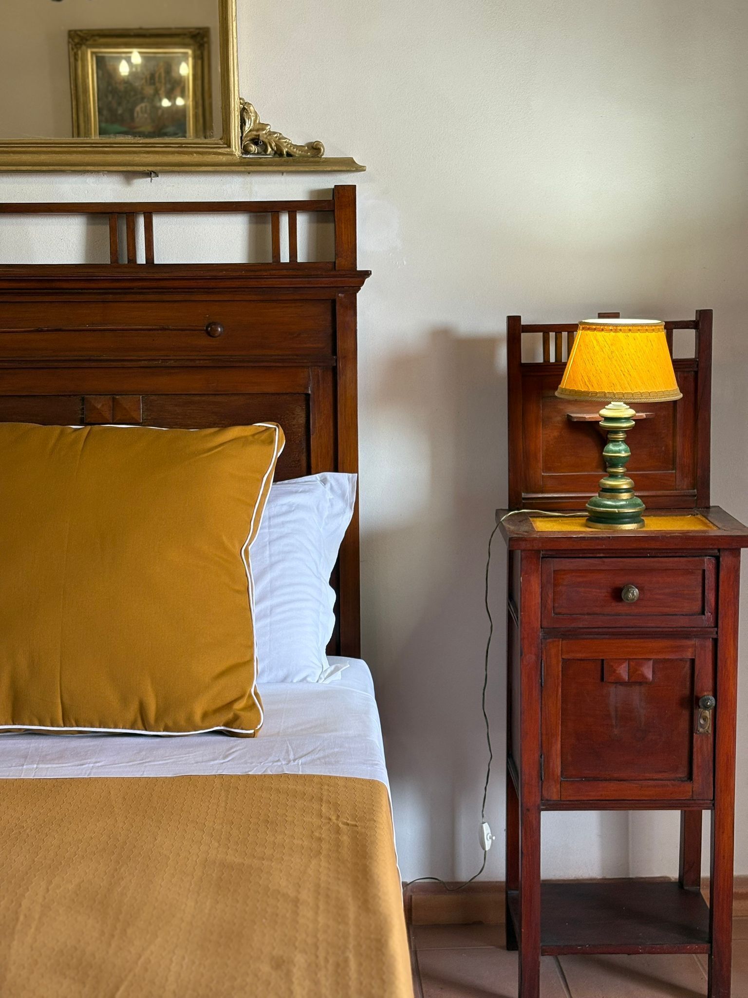 Una cama con cabecera de madera, un almohadón amarillo y un almohadón blanco. Junto a la cama, una mesita de noche de madera con una lámpara y un cajón. En la pared, encima de la cama, cuelga un cuadro enmarcado.