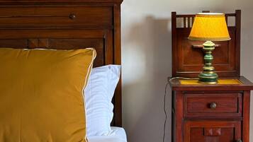 Una cama con cabecera de madera, un almohadón amarillo y un almohadón blanco. Junto a la cama, una mesita de noche de madera con una lámpara y un cajón. En la pared, encima de la cama, cuelga un cuadro enmarcado.