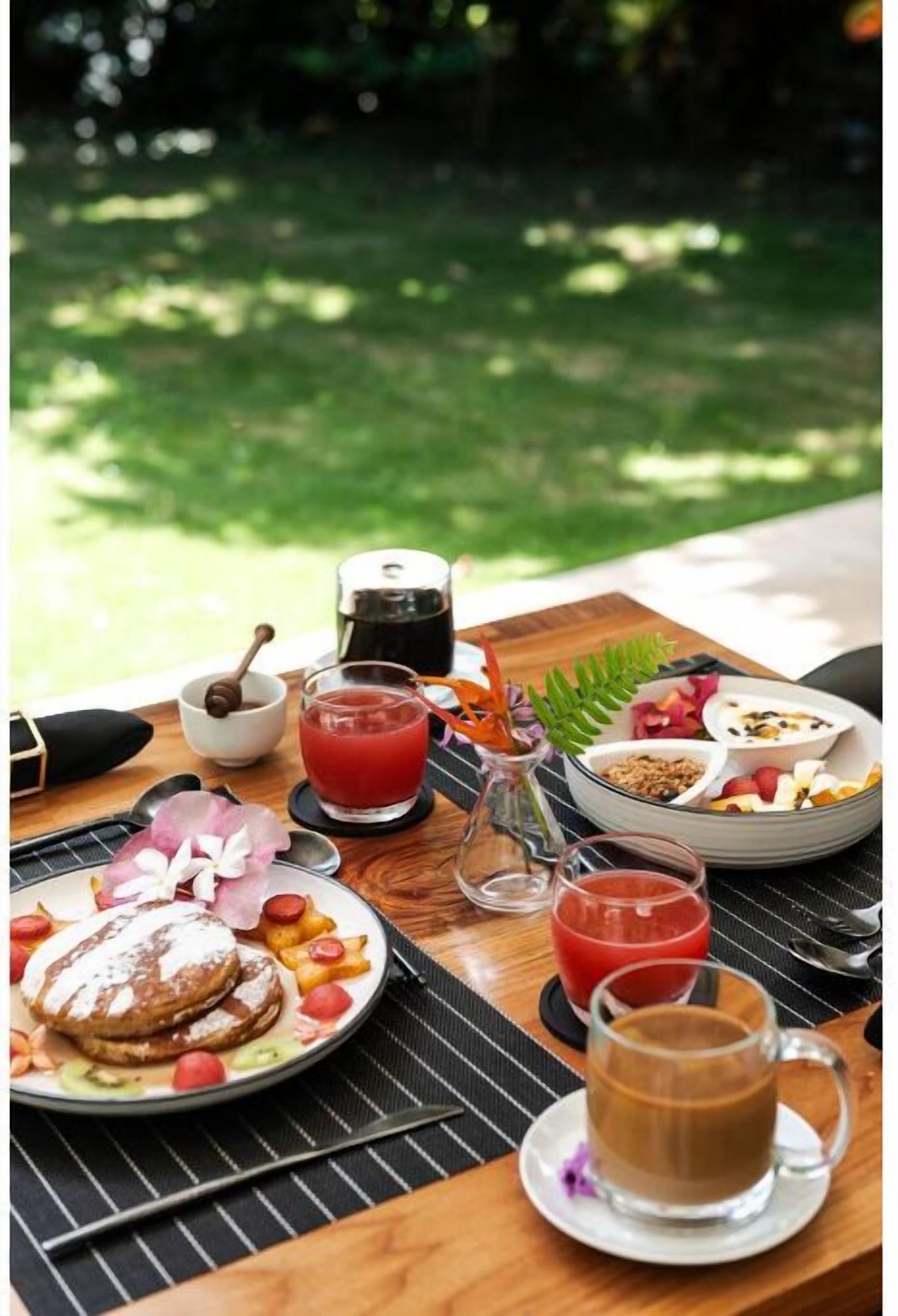 Une table de petit-déjeuner avec des crêpes, des fruits et du café, installée à l’extérieur, offrant une vue sur la verdure.