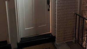 A white door with a small window and a peephole, a doormat with "STAY AWAY" text, and a brick wall with a light fixture. - Cozy Home, 17 min from NYC, 2 Parking Spaces (Ridgefield Park)