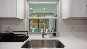 A kitchen with a stainless steel sink, a window overlooking a backyard with a pool and chairs, and a small potted plant on the windowsill. - Cozy Pool House 24 min to Beach, Cozy Pool House 24 min to Beach (Manatee County)