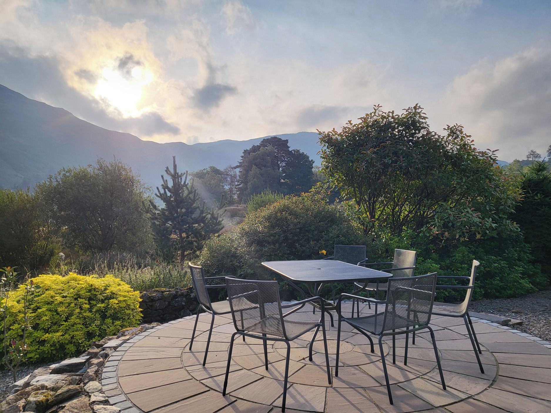An outdoor patio with a table and chairs, surrounded by greenery and mountains in the background.