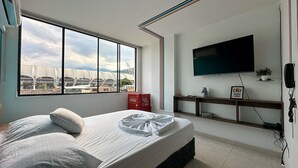 A bedroom with a large window, a bed, a television, and a shelf with a plant and a sign. - San fernando 405 estadio a una cuadra (Cali)