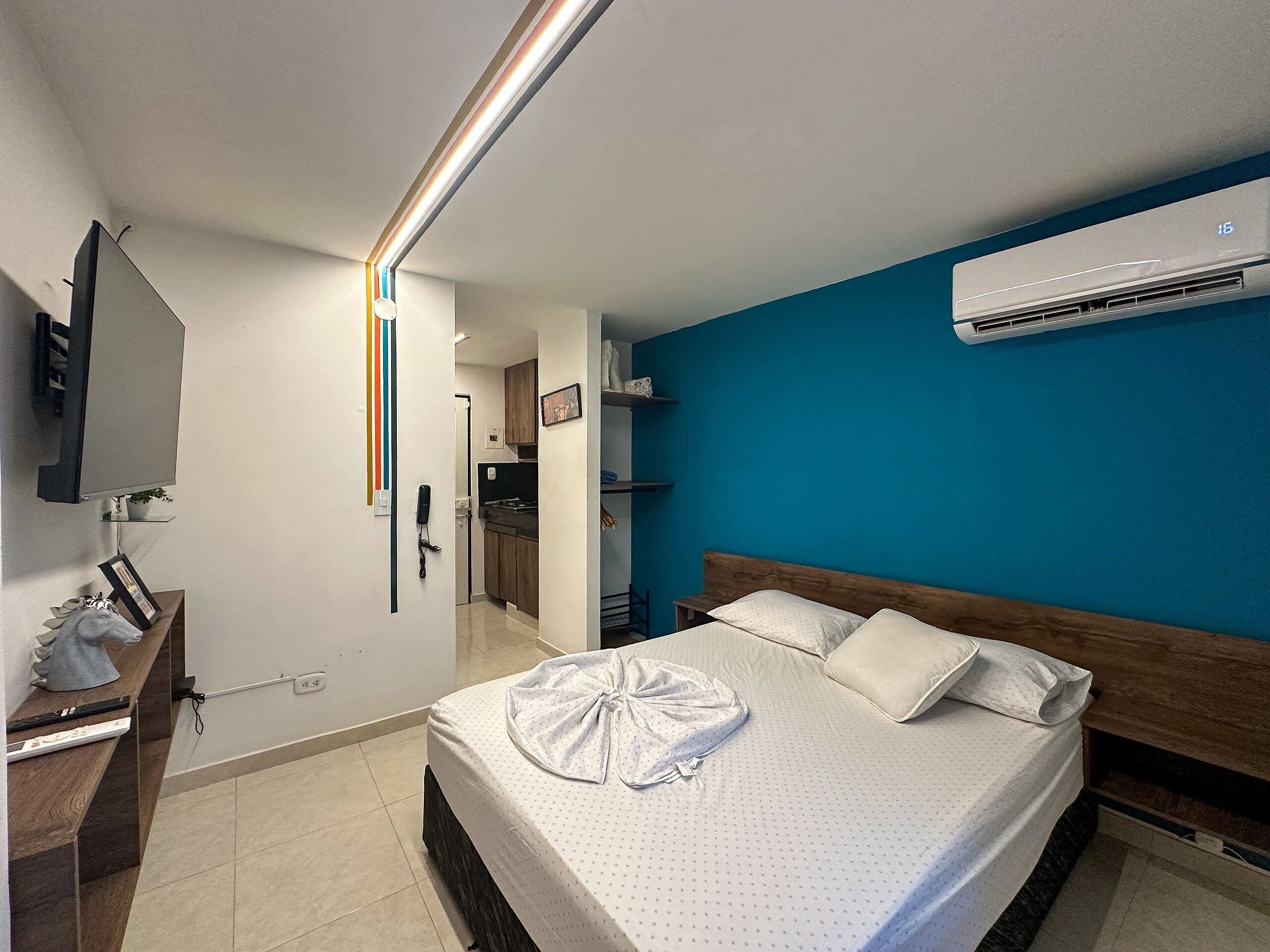 A hotel room with a bed, a TV, an air conditioner, a desk with a lamp, and a bathroom visible through an open door.