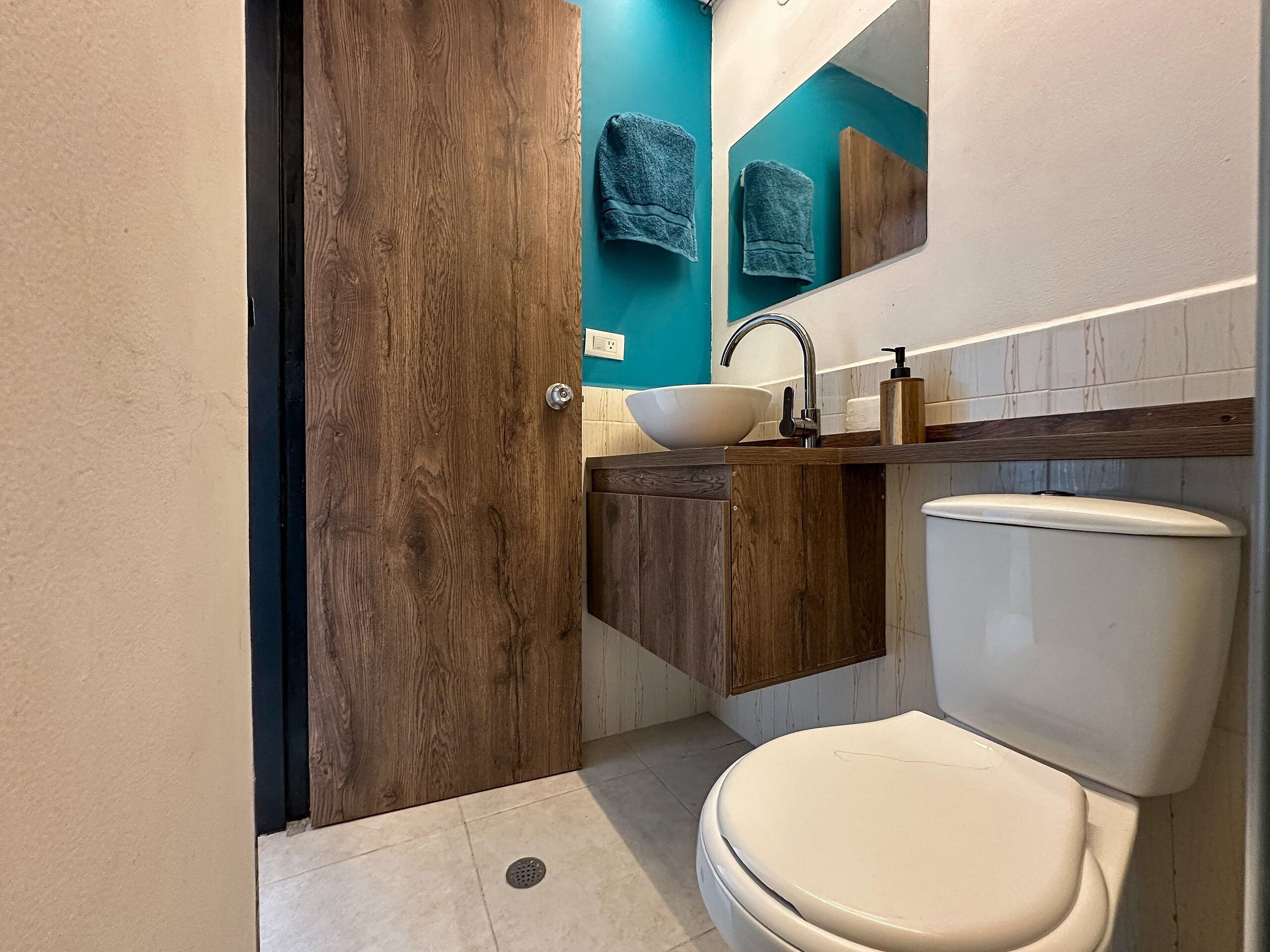 Une salle de bain avec une porte en bois, un lavabo blanc, des toilettes blanches et un miroir au-dessus du lavabo.
