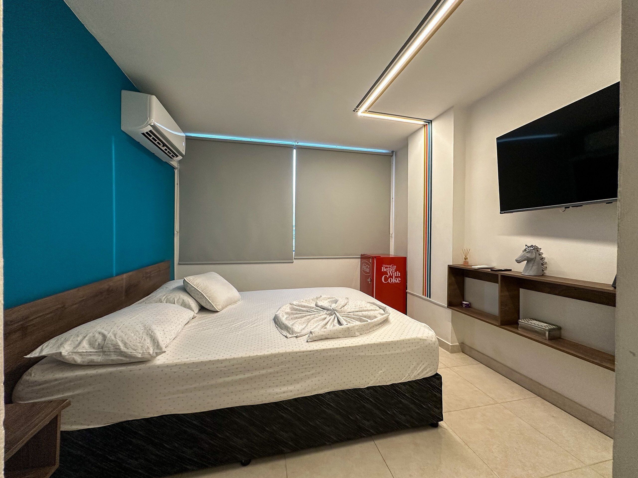 A modern hotel room with a bed, a mounted TV, a shelf with decorative items, and a window with blinds.