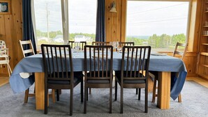 Un coin repas avec une table dressée pour quatre personnes, entouré de chaises en bois, de grandes fenêtres avec des rideaux et une vue sur un paysage rural.