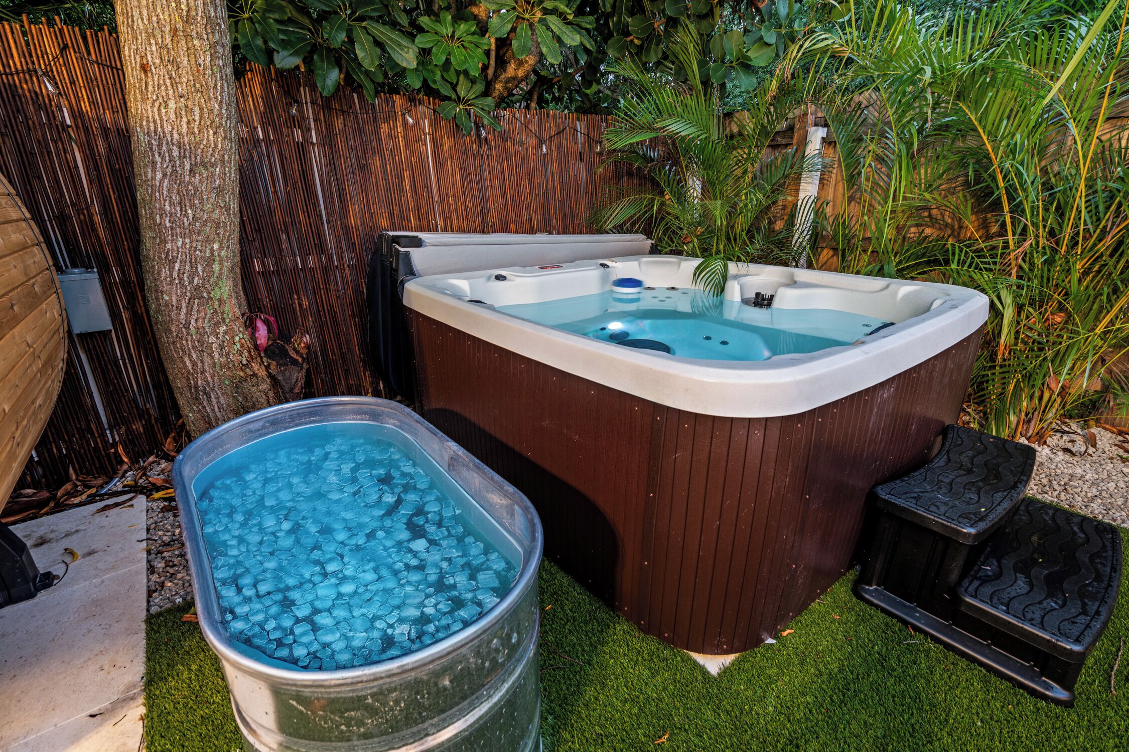 A hot tub with a wooden exterior, surrounded by a bamboo fence and lush greenery.