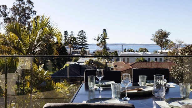 A dining table set for four with clear glasses and plates, overlooking a coastal view with buildings and trees. - The Palms-  9 Pantowora St, Corlette (Corlette)