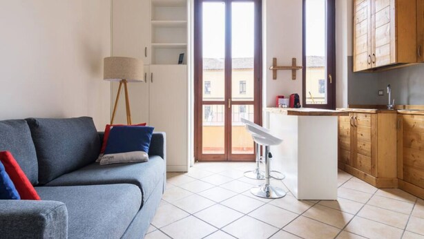 A modern kitchen with wooden cabinets, a built-in island, and a dining area with a bar stool. - Cozy apartment in Bovisa (Milano)