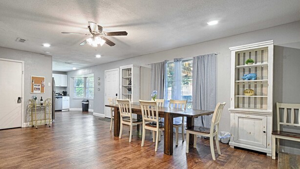 Un grand espace à manger avec une table et des chaises en bois, un ventilateur de plafond et une cuisine en arrière-plan.