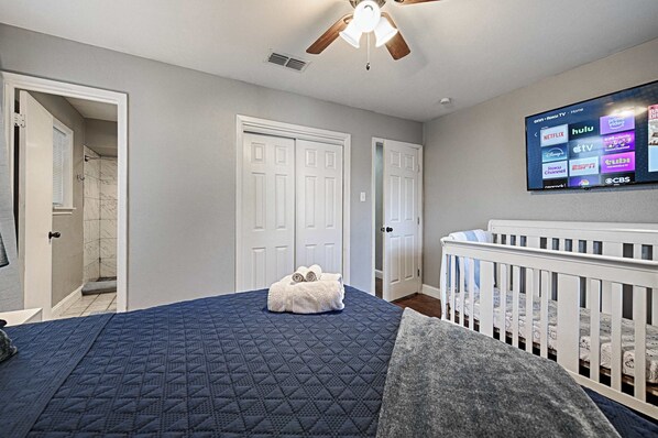 A bedroom with a bed, a crib, a ceiling fan, and a television displaying streaming services.