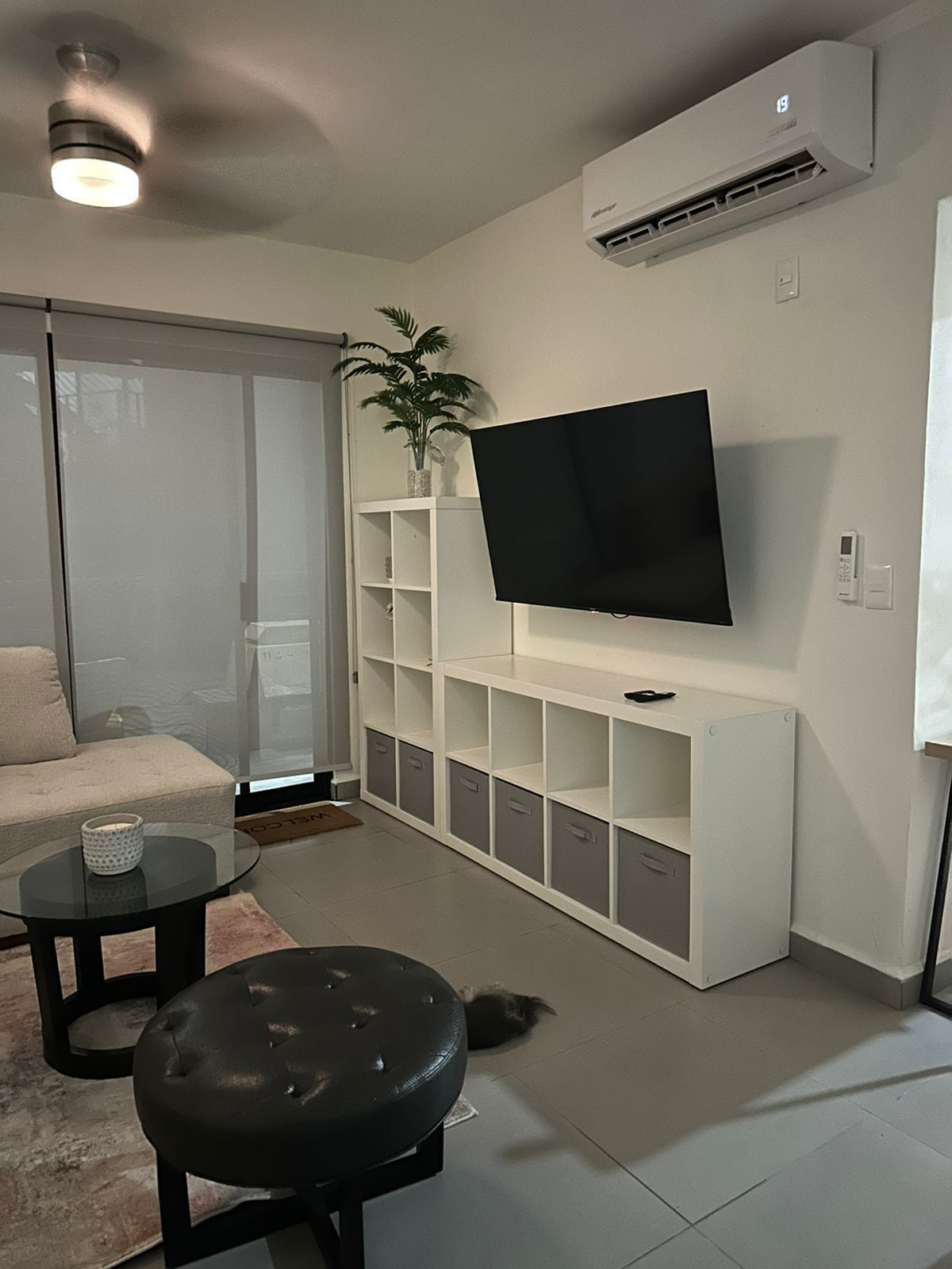 Een moderne woonkamer met een flatscreen televisie bevestigd aan een wit kastje, een glazen salontafel, een zwart poefje, een beige bank en een potplant.