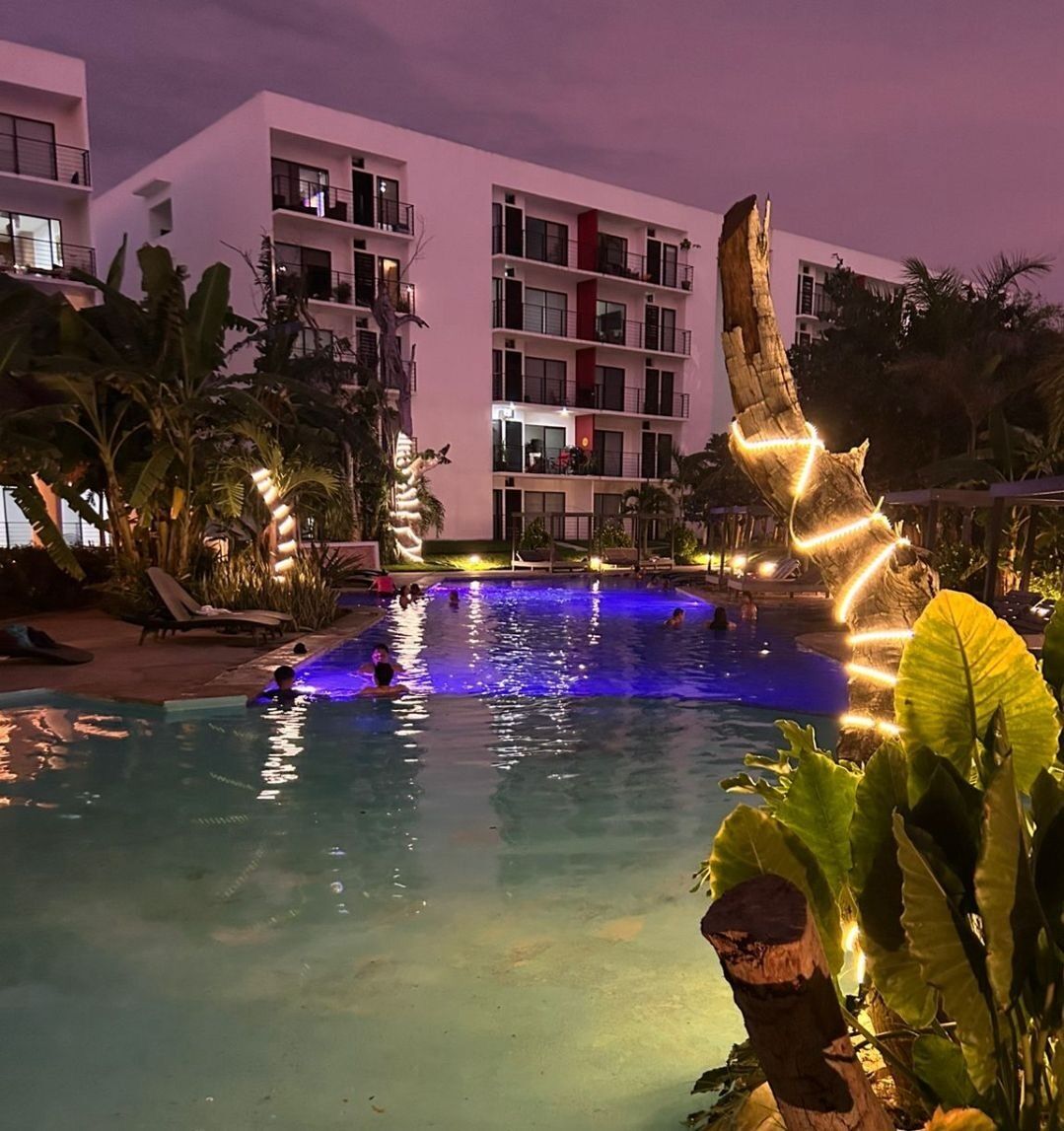 A swimming pool with illuminated edges, surrounded by palm trees and a multi-story building with balconies.