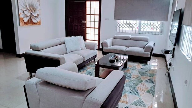 Ein Wohnzimmer mit Sofa, Sessel und Couchtisch. Eine Tür, ein Fenster mit Jalousien und ein Bild an der Wand sind ebenfalls vorhanden.