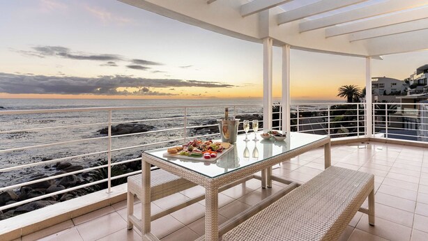 A balcony with a glass table set with food and drinks, overlooking a beach. - Sunset Azure Penthouse - Panoramic Views, PVT Pool (Cape Town)