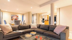 A modern living room with a sectional sofa, a glass coffee table, and a dining area with a kitchen in the background. - Sunset Azure Penthouse - Panoramic Views, PVT Pool (Cape Town)