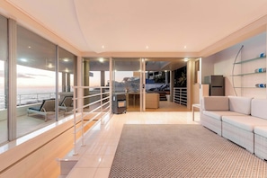 A modern living room with a sofa, a patterned rug, a kitchen area, and a view of the sea through large windows.