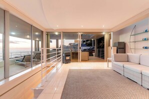 A modern living room with a sofa, a patterned rug, a kitchen area, and a view of the sea through large windows. - Sunset Azure Villa - Sea Views, Sleeps 14, 2 Pools (Cape Town)